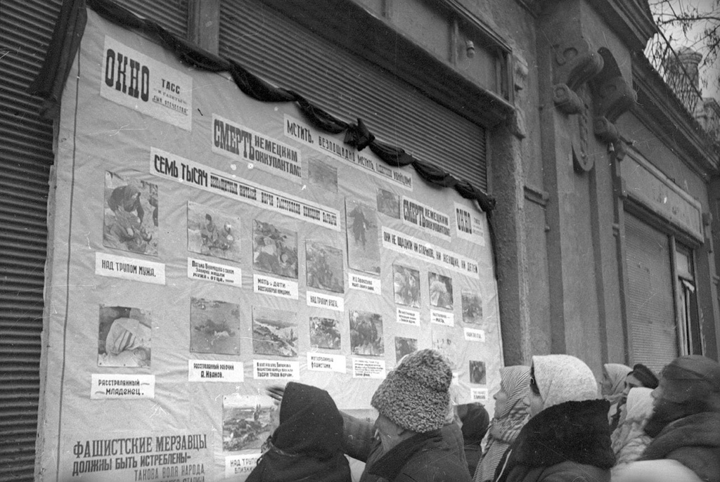 Окна ТАСС. Керчане у стенда с фотографиями жертв расстрела в Багеровском рву, 1942 год, Крымская АССР, г. Керчь. В течение первых дней декабря 1941 года несколько тысяч (согласно немецкому рапорту - 2,5 тысячи человек, согласно свидетельствам очевидцев - 7 тысяч человек) евреев, живших в Керчи, были расстреляны зондеркомандой 10Б айнзатцгруппы Д в противотанковом рву у поселка Багерово. Затем, 29 декабря, в том же противотанковом рву в связи с тем, что советской разведгруппой был убит немецкий офицер, оккупанты расстреляли несколько сотен жителей поселка Самострой около Камыш-Буруна - русских, украинцев, крымских татар.