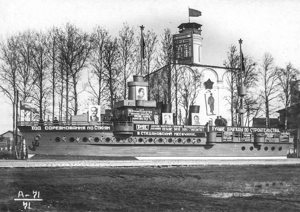 Вид трибуны на первом участке, 1937 год, Северная обл., пос. Судострой. Октябрьская площадь поселка Судострой. Как преобразился Никольский собор! На его центральном барабане развевается флаг. Барабан  частично закрыт стендом «Стахановский месячник 20 марта – 20 апреля». Северная стена собора украшена пятиконечной звездой и ростовым портретом Сталина. Перед собором выстроена трибуна в виде корабля. На «носу» трибуны-корабля флажок «9 апреля 1937 года». Трибуна украшена портретами членов правительства и транспарантами «Ход соревнования по строям», «Привет рабочим, инженерам, служащим Водоканалстроя, получившим переходящее Красное знамя строительства», «Лучшие бригады по строительству». На трибуне стоят люди.Рабочий поселок Судострой в 1938 году получил статус города и название Молотовск. В 1957 году переименован в Северодвинск. 
