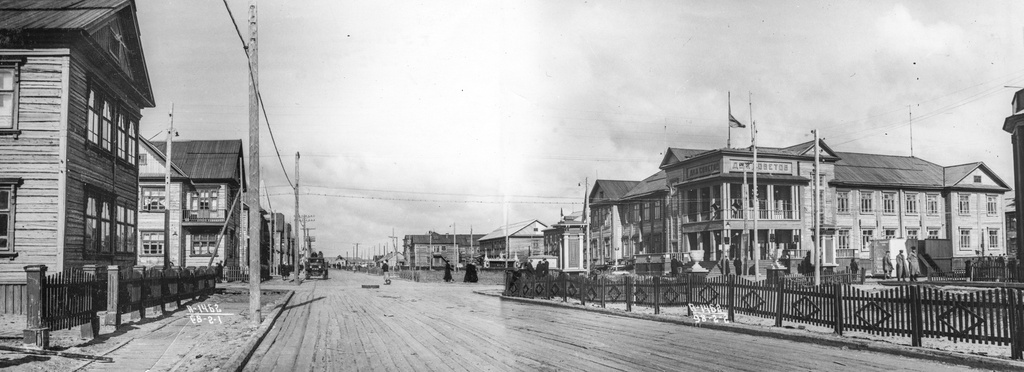 Вид со стороны Советской улицы на Дом Советов, 1 июня 1940, Архангельская обл., г. Молотовск. Пересечение двух главных улиц города – Советской и проспекта Беломорского.  В центре здание Дома Советов, здесь располагалась горком и горисполком Молотовска, а сейчас находится горвоенкомат. Большое крыльцо и пристроенный сбоку балкон служили трибуной во время городских праздников. Над домом развивается флаг Советского Союза, а на балконе второго этажа видны раструбы громкоговорителей. Улица Советская разделена на две части, с одной стороны идет деревянная дорога, с другой – небольшой бульвар с клумбами и скамейками, огороженный штакетником.Город Молотовск в 1957 году переименован в Северодвинск. 