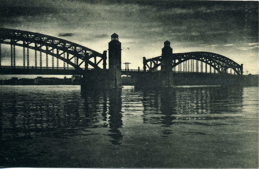 Охтенский мост (№ 31), 1920 - 1930, г. Ленинград. Мост Императора Петра Великого (Большой Охтенский мост) – Большеохтинский мост.1911–1917 – Мост Императора Петра Великого;1917–1956 – Большеохтенский мост;С 1956 – до настоящего времени – Большой Охтенский мост илиБольшеохтинский мост.Авторы: инженер-полковник Григорий Григорьевич Кривошеин, инженер-полковник Владимир Петрович Апышков, в доработке проекта принимали участие инженер-полковник Сергей Павлович Бобровский, инженер-технолог П. М. Шелоумов, инженер путей сообщения Григорий Петрович Передерий.По проекту инженера-полковника Г. Г. Кривошеиным мост имеет в длину 160 саж. и в ширину 11 саж., при трех пролетах и двух быках; средний пролет в 20 саж. – подъемный и боковые по 60,15 саж. в ширину и 4,15 саж. в высоту – перекрыты двумя двухшарнирными арочными фермами с затяжкою и ездою по низу.Каждая ферма состоит из двух арочных поясов, соединенных между собою раскосами и стойками. К нижнему поясу прикреплены на расстоянии 3,7 саж. подвески, а к ним затяжки и поперечные балки, на которых покоится проезжая часть. Высота ферм посредине достигает 22,6 метра.Быки предположено было основать на кессонах, опущенных в грунт на глубину 11–12 1/2 саж. Тело быка предлагалось облицевать гранитом: серым до ординара воды и красным выше. Ледорезы одинаковые, по типу ледорезов Троицкого моста, с несколько улучшенной разрезкою. Над ледорезами предлагалось поместить головы Нептуна.На быках с передней части моста находиться по одной башне для помещения разводного механизма, а наверху фонаря маяка. Тело башен предположено из бетона на гравии, с гранитной облицовкой. Форма башни 6-гранная с закругленными углами. Сбоку башни примыкает полубашня, назначенная для механика и для размещения всех приводов для управления разводкой.Разводной пролет проектирован в виде раскрывающегося подъемного моста в два крыла.Механическое оборудование предположено электрогидравлическое.Торжественная закладка моста произошла накануне дня 200-летия Полтавской битвы – 26 июня 1909 года.Мост решено было назвать в честь триумфатора этой битвы – императора Петра Великого – Охтенский Императора Петра Великого мост.26 октября 1911 года был произведен торжественный пуск моста в эксплуатацию в присутствии министра внутренних дел А. А. Макарова, хотя строительные работы еще продолжались до 1913 года.Ленинград с августа&nbsp;с&nbsp; августа 1914 года до 26 января 1924 года – Петроград.&nbsp;Фотооткрытка с фотографии фотографии В. В. Преснякова.