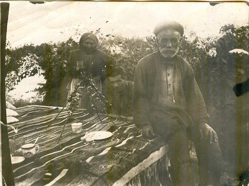 Отец Саркиса и его бабушка в саду за приготовлением шашлыка, 1955 - 1957, Армянская ССР, г. Дилижан. На первом плане перед столом Григор Багдасарян. За столом сидит Саркис Багдасарян, рядом с ним бабушка.