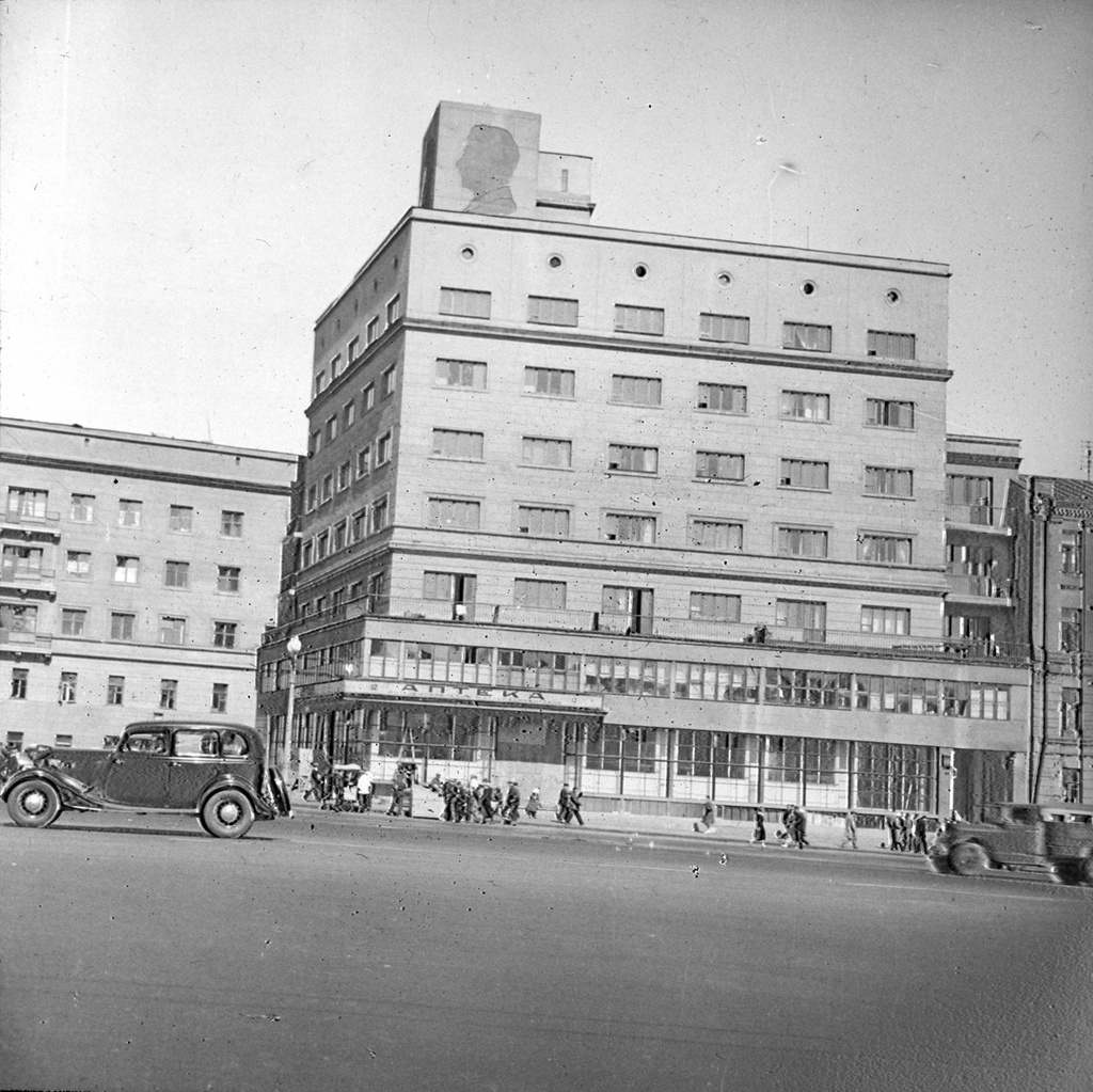 На улице города, 16 апреля 1956 - 14 мая 1956. На здании сверху был барельеф Сталина. В рамках борьбы с культом личности, остался только контур.Фотография сделана британской делегацией, побывавшей в СССР в 1956 году.Выставка «Английские энергетики в Советском Союзе» с этим снимком.