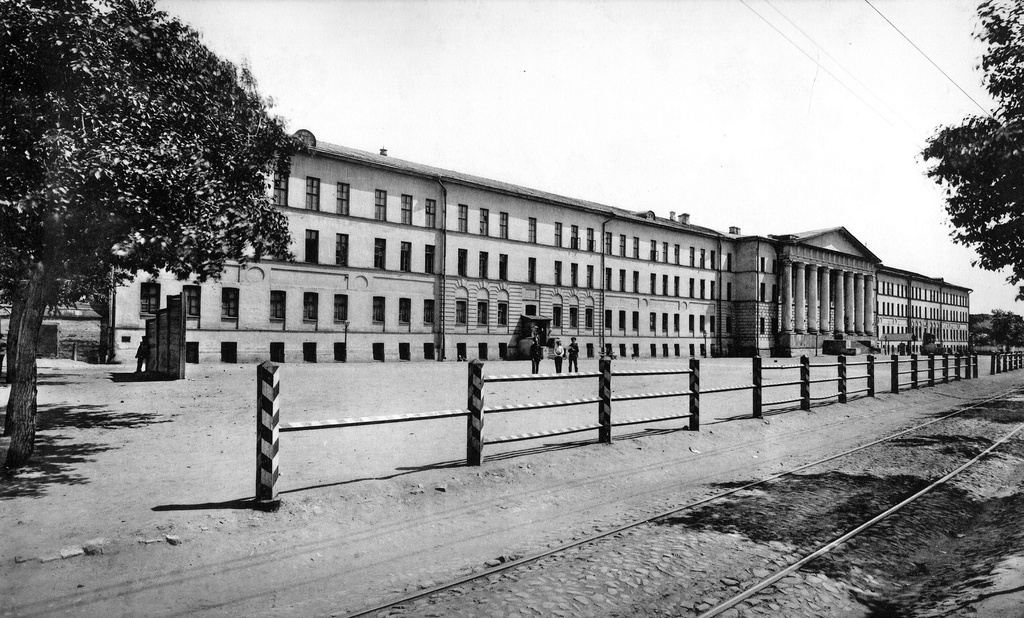 Покровские казармы, плац перед казармами и трамвайная линия маршрута "А", 1916 - 1920, г. Москва. Покровские казармы – детище Павла I, предложившего московскому населению освобождение от постойной повинности в обмен на добровольное финансирование строительства военных казарм. Постойную повинность несли все собственники жилья, кроме членов императорской фамилии, иностранных дипломатов и высших церковных иерархов. На такое строительство требовалось 2.5 млн.руб. Стараниями москвичей собрали около полумиллиона. На дома тех, кто дал деньги, прикреплялась табличка «Свободен от постоя». Такие таблички до сих пор можно видеть на некоторых старинных зданиях. В 1798–1801 годах был выстроен единственный корпус для мушкетёрного батальона (семь рот Навашинского полка) у Покровских ворот. В 1812 году казармы горели. В 1830-х годах архитекторы И. Жилярди и Ф. Соколов перестроили казармы – возвели третий этаж и более чем вдвое расширили здание. Здание приобрело монументальный фасад с мощным восьмиколонным портиком, увенчанным массивным фронтоном с барельефом двуглавого орла. Позднее внутри казарм, в правом крыле, была устроена церковь Осии Пророка в память чудесного спасения Императорской семьи 17 октября 1888 года. Эта церковь служила полковым храмом 7-го гренадерского Самогитского полка. Церковь была закрыта в 1919 году. В 1930-х годах казармы надстроены четвертым этажом. Орла на фронтоне сменил герб СССР. Перед казармами на месте нынешнего Покровского бульвара находился обширный плац. В 1891 году плац был уменьшен и огражден. Вдоль плаца проходили линии сначала конки, а с 1911 года — трамвая. В 1954 году плац был упразднен, вдоль казарм открыт проезд для транспорта, трамвайные линии стали проходить по обе стороны устроенного здесь бульвара. В казармах сначала квартировали мушкетеры, затем 7-й Гренадерский Самогитский полк, с 1904 года 3 и 4-й батальоны лейб-гренадерского Екатеринославского полка, позднее 56-й запасный полк. В 1933–1960 годах казармы носили имя Ф.Э. Дзержинского. В мае 1960 года военная история казарм закончилась – здание передали Госснабу, сегодня его занимают организации нефтегазовой отрасли.