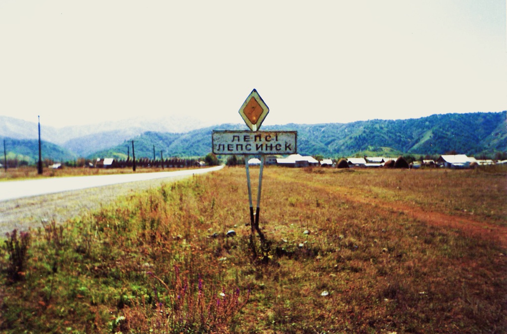 Въезд в Лепсинск, 1990 год, Казахская ССР, Талды-Курганская обл., Саркандский р-н, с. Лепсинск. На авторском снимке  на фоне гор изображен въезд в Лепсинск, с дорожным знаком с вывеской на казахском и русском языках «Лепсi» – «Лепсинск». Казачья станица Лепсинская, или Верх-Лепсинская, была образована сибирскими казаками и крестьянами в 1855 году в горной котловине Чубар-Агач, в Джунгарском Алатау.