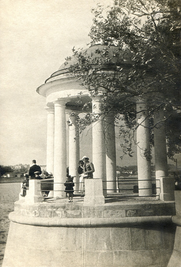Портрет жены и сына в беседке, 1936 год, г. Москва. Семья автора на отдыхе: жена (в белой шляпке), рядом, слева у перил, стоит сын (4 года) в пилотке. Беседка ЦПКиО имени Горького на набережной Москвы-реки. Автор – Борис Александрович Баш (Шишлянников) (1901–1942), фотограф, кинооператор и режиссер.