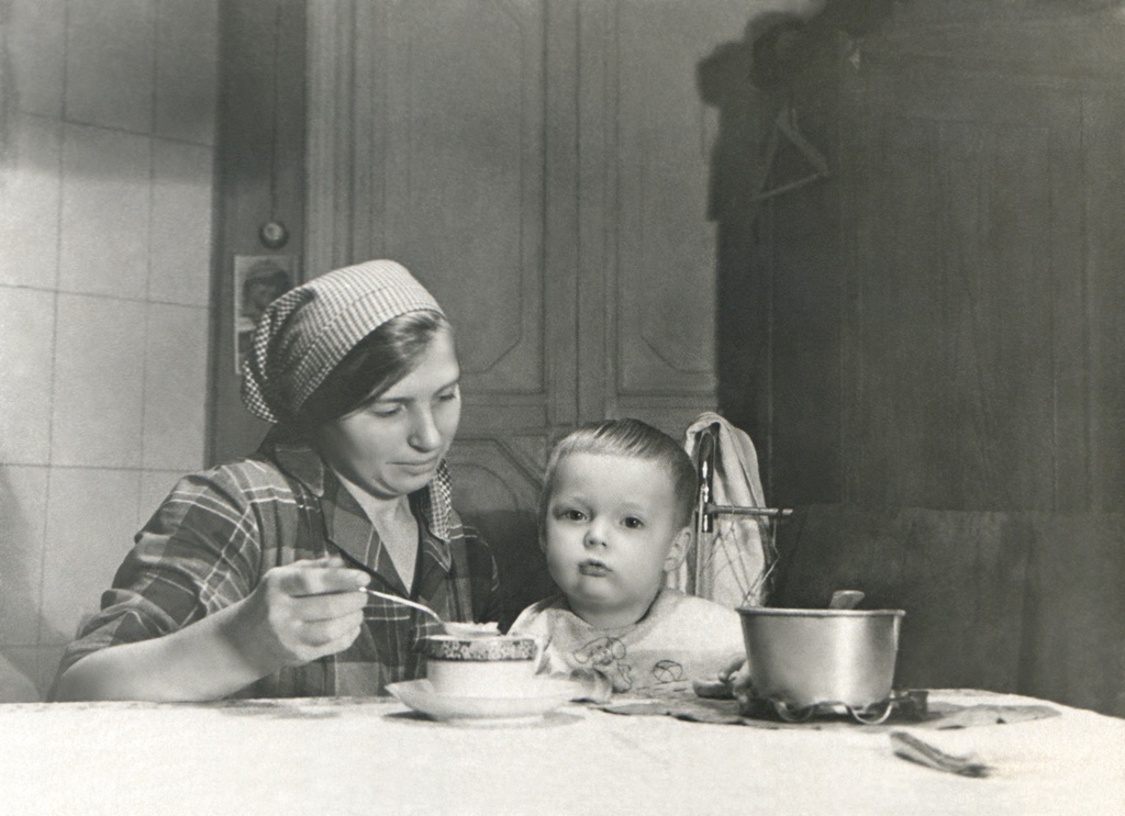 Портрет сына (2 года) и домработницы Лиды в комнате, 1934 год, г. Москва. Комната на верхнем этаже в коммунальной квартире, метров 15-20, где жил автор с женой, сыном и тещей. Справа – детская металлическая постель с ограждающей сеткой и шкаф. Слева от двери: выключатель (открытая проводка), под ним висит отрывной календарь. Один из признаков коммунальной квартиры на любом фото – супница или просто кастрюля на столе, которую приносили в комнату с коммунальной кухни. Почти все фото на Житной улице, дом 10 сделаны в этой комнате. Автор – Борис Александрович Баш (Шишлянников) (1901–1942), фотограф, кинооператор и режиссер.