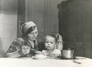 Портрет сына (2 года) и домработницы Лиды в комнате, 1934 год, г. Москва. Комната на верхнем этаже в коммунальной квартире, метров 15-20, где жил автор с женой, сыном и тещей. Справа – детская металлическая постель с ограждающей сеткой и шкаф. Слева от двери: выключатель (открытая проводка), под ним висит отрывной календарь. Один из признаков коммунальной квартиры на любом фото – супница или просто кастрюля на столе, которую приносили в комнату с коммунальной кухни. Почти все фото на Житной улице, дом 10 сделаны в этой комнате. Автор – Борис Александрович Баш (Шишлянников) (1901–1942), фотограф, кинооператор и режиссер.