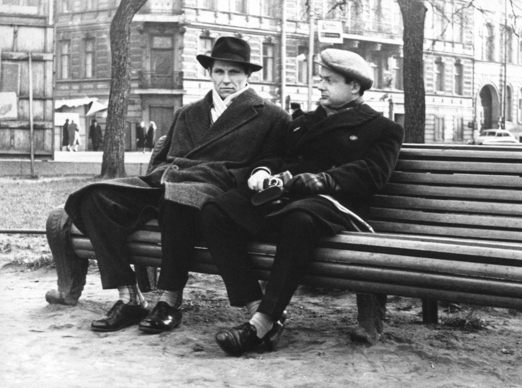 В сквере на Манежной площади, 1961 год, г. Ленинград. Слева – коллега, справа – Олег Борисович Баш, у него в руках фотоаппарат «Смена-6» с чехлом, который начал выпускаться с 1961 года. На фото два дома: угол дома 37/18 на Итальянской улице, правее – Караванная улица, дом 20 (нумерация современная). За скамейкой, на столбе висит указатель маршрутов на остановке общественного транспорта.