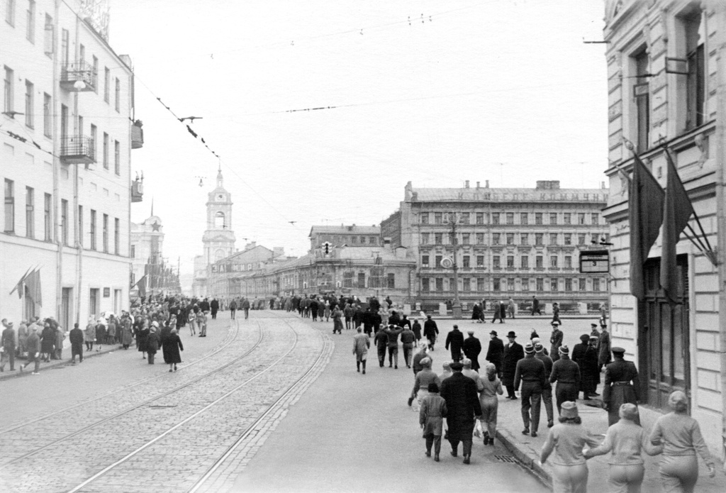 Улица Балчуг, 1961 - 1963, г. Москва. Вероятно, фото сделано из кузова грузовой машины. Прямо – Чугунный мост и начало Пятницкой улицы. Слева на тротуаре – продавщица в белом халате, к ней очередь, уходящая на мост. Справа: люди идут с первомайской демонстрации, среди них – девушки-физкультурницы. На доме – указатель остановки трамвая 3-го маршрута.