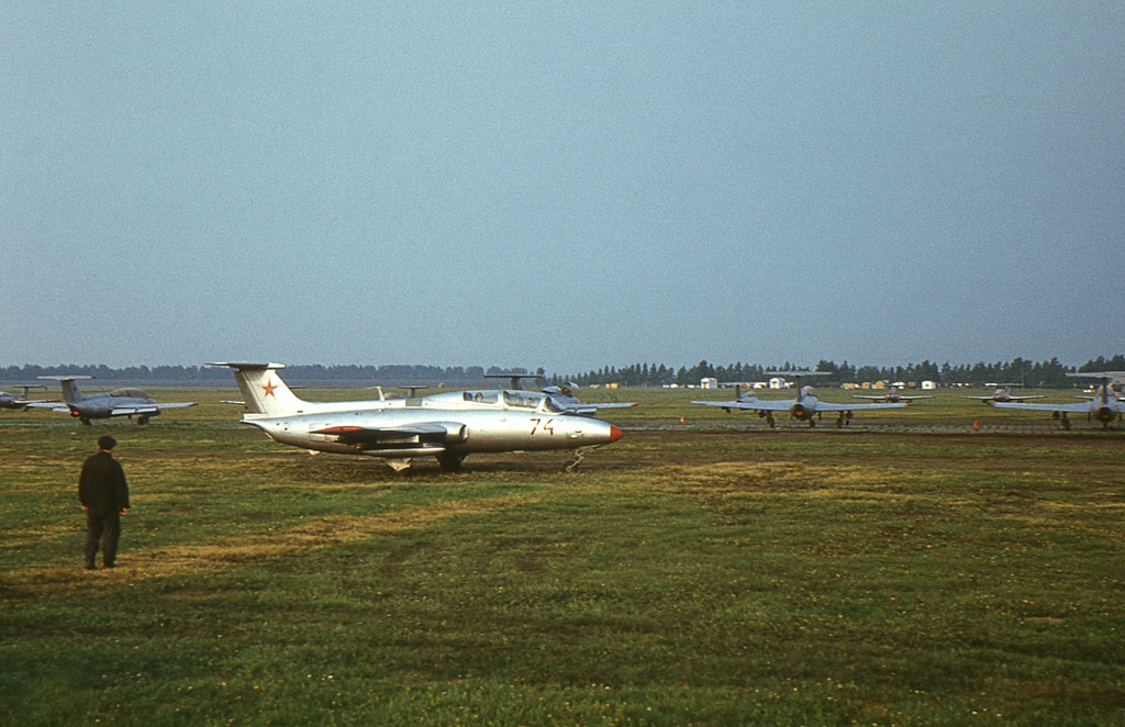Полеты на грунтовом аэродроме в Кирсанове, 643 уап ТВВАУЛ, июнь - август 1982, Тамбовская обл., Кирсановский муниципальный округ, Голынщинский сельсовет. Аэродром в городе Кирсанове. Борт № 74 заруливает на стоянку из «железки». Слева, в синей форме, стоит техник самолета или кто-то из тех. состава по обслуживанию самолетов.На дальнем плане, у лесополосы, маленькие белые и бежевые домики – служебные помещения каждой эскадрильи. Казармы располагались на краю Кирсанова, в районе Приовражной улицы (справа за кадром).643 учебный авиаполк ТВВАУЛ (в/ч 42149, база: город Тула, аэродром Клоково, самолеты Л-29). На лето авиаполк перелетал из Тулы в Кирсанов и Новгородовку Тамбовской области (по две эскадрильи на каждый город).ТВВАУЛ – Тамбовское высшее военное авиационное училище летчиков имени М. М. Расковой.Часть расформирована в 1990 году, потом в 1995 году закрыли и Тамбовское ВВАУЛ.