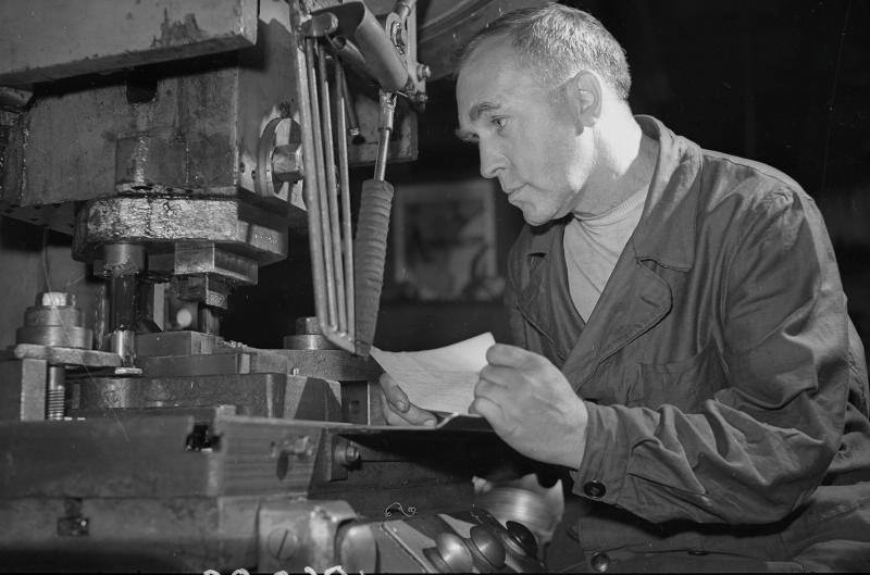 Рабочий у станка, 1955 - 1965. Рабочий держит небольшой лист бумаги и смотрит на станок.