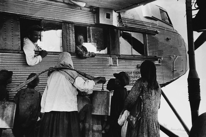 Агит-эскадрилья имени Горького. Остановка в пути, 1938 год. Из серии «Парашютный отряд агитэскадрильи „Максим Горький”».Сбор средств на создание агитэскадрильи имени Максима Горького начался в октябре 1932 года по инициативе журнала «Огонек» в честь 40-летнего юбилея творческой деятельности Максима Горького. Приказом от 17 марта 1933 года объявлено формирование агитэскадрильи, с 5 мая (День Печати) 1933 года началась ее регулярная работа.